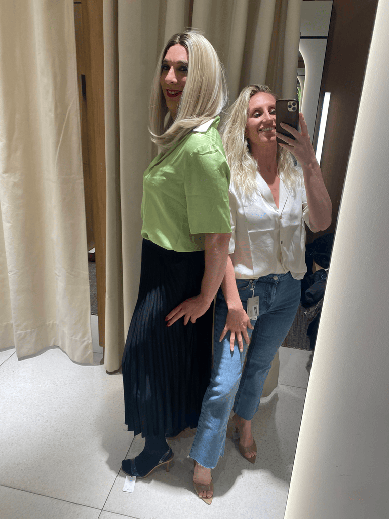 Photo de Jennifer, maquilleuse professionnelle, prenant un selfie avec Stella, une de ses clientes transgenres, lors d'une séance shopping. Jennifer, souriante, porte une chemise blanche et un jean, tandis que Stella arbore une blouse verte à manches courtes et une jupe plissée noire, reflétant le style et la confiance acquis grâce à l'accompagnement personnalisé de Jennifer.