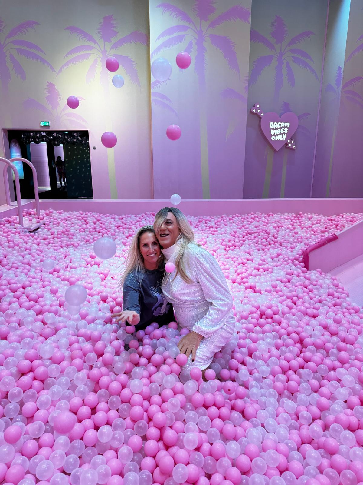 Jennifer et Deborah profitent d'un moment de détente et de fun dans une piscine de balles roses, sous un décor pastel à l'ambiance rêveuse. Ce cadre inspirant de l'exposition 'House of Dreamers' reflète leur joie et leur complicité, symbolisant la liberté d'exprimer leur féminité avec confiance