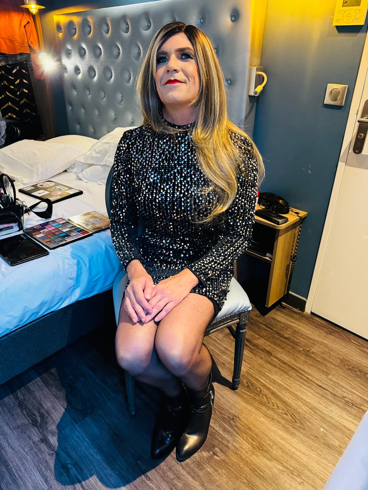 Stéphanie, vêtue d'une robe noire à sequins, est assise dans une chambre d'hôtel avec un sourire doux. Ses mains sont posées sur ses genoux, et son regard exprime calme et sérénité