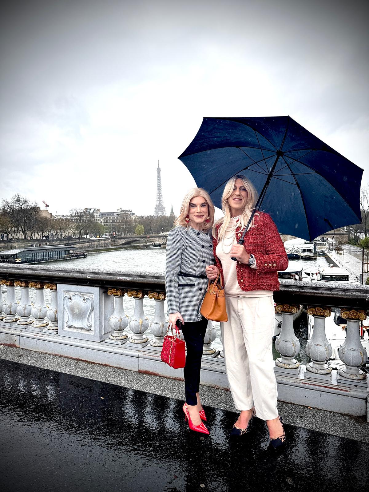 Freda à gauche et Queen D à droite sous un parapluie à Paris, avec la tour Eiffel en fond, lors d’une sortie Transbeauté entre féminisation et accompagnement travesti