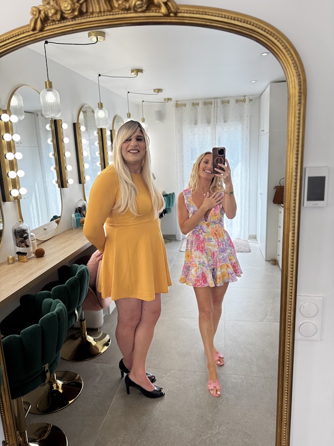 Deux personnes en robe posent devant un miroir doré au studio Transbeauté, l’une en jaune moutarde, l’autre en robe fleurie.